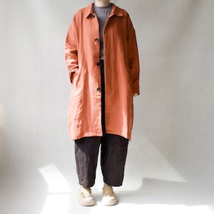 Könnte beinhalten: Ein langer, terracottafarbener Leinenmantel mit Knopfleiste und lockerer Passform. Der Mantel wird über einem dunkelgrauen T-Shirt und einer dunkelgrauen Hose getragen.
