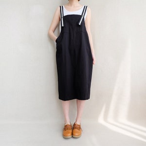 May include: A black midi-length overall dress with white straps. The straps have a white stripe down the center. The dress has a relaxed fit and is made of a soft, comfortable fabric.