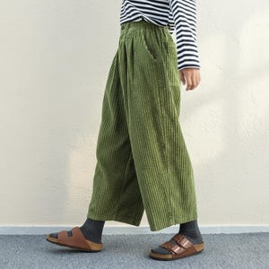 May include: Olive green corduroy pants with a wide leg design. The pants have a high waist and pockets. The person is wearing a black and white striped long-sleeve shirt and brown sandals.