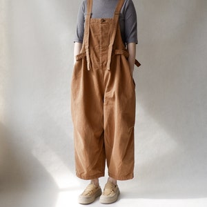 May include: A brown overall jumpsuit with wide legs and adjustable straps. The straps are tied at the waist. The jumpsuit is made of a thick, textured fabric. The model is wearing the jumpsuit with a grey short-sleeved shirt and brown leather shoes.