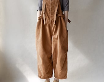 May include: A brown overall jumpsuit with wide legs and adjustable straps. The straps are tied at the waist. The jumpsuit is made of a thick, textured fabric. The model is wearing the jumpsuit with a gray short-sleeved shirt and brown leather shoes.