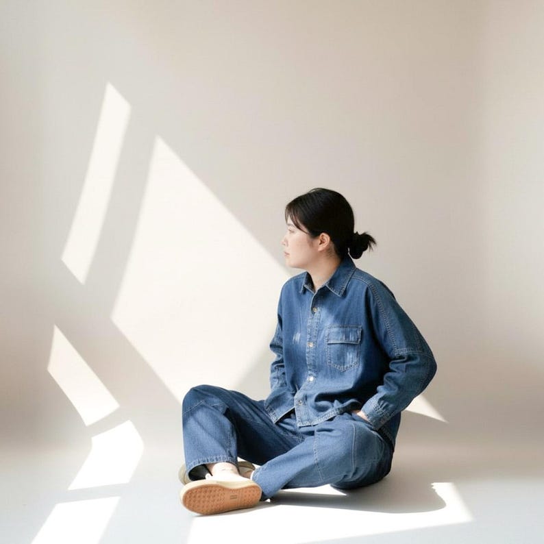 May include: A denim shirt and matching pants set in a medium wash. The person is sitting on the floor with their legs crossed, wearing beige shoes. The background is a plain white wall with shadows.