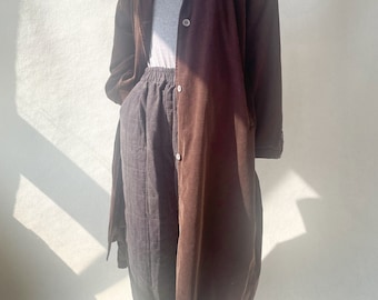 May include: A long brown corduroy coat with a button closure and a relaxed fit. The coat is worn over a gray t-shirt and black pants.