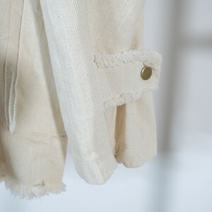 Op de afbeelding: Close-up van de manchet van een wit denim jack met een rafelige rand en een gouden knoop.