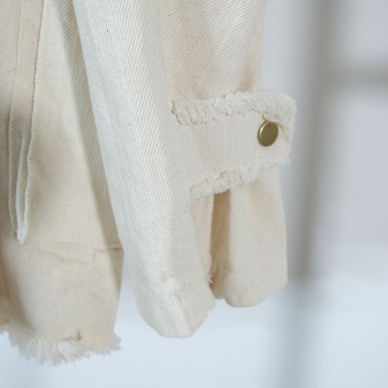 Op de afbeelding: Close-up van de manchet van een wit denim jack met een rafelige rand en een gouden knoop.