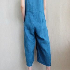 May include: A teal blue linen overall jumpsuit with wide legs. The jumpsuit has thin straps and a square neckline. The model is wearing the jumpsuit with a white tank top underneath.