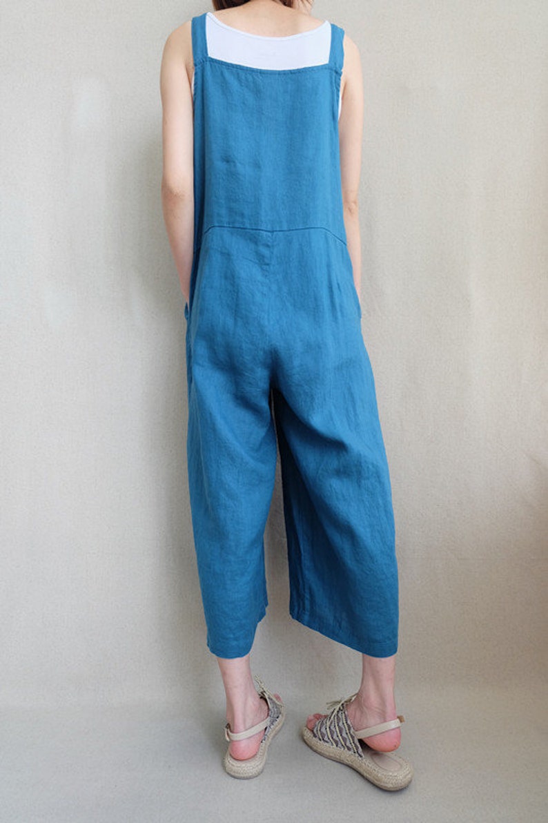 May include: A teal blue linen overall jumpsuit with wide legs. The jumpsuit has thin straps and a square neckline. The model is wearing the jumpsuit with a white tank top underneath.