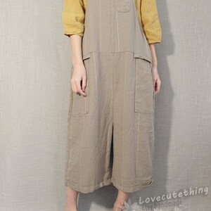 May include: A light brown linen overall dress with thin white stripes. The dress has two large pockets on the front and a smaller pocket on the chest. The model is wearing a yellow short-sleeved shirt under the dress and brown leather shoes.