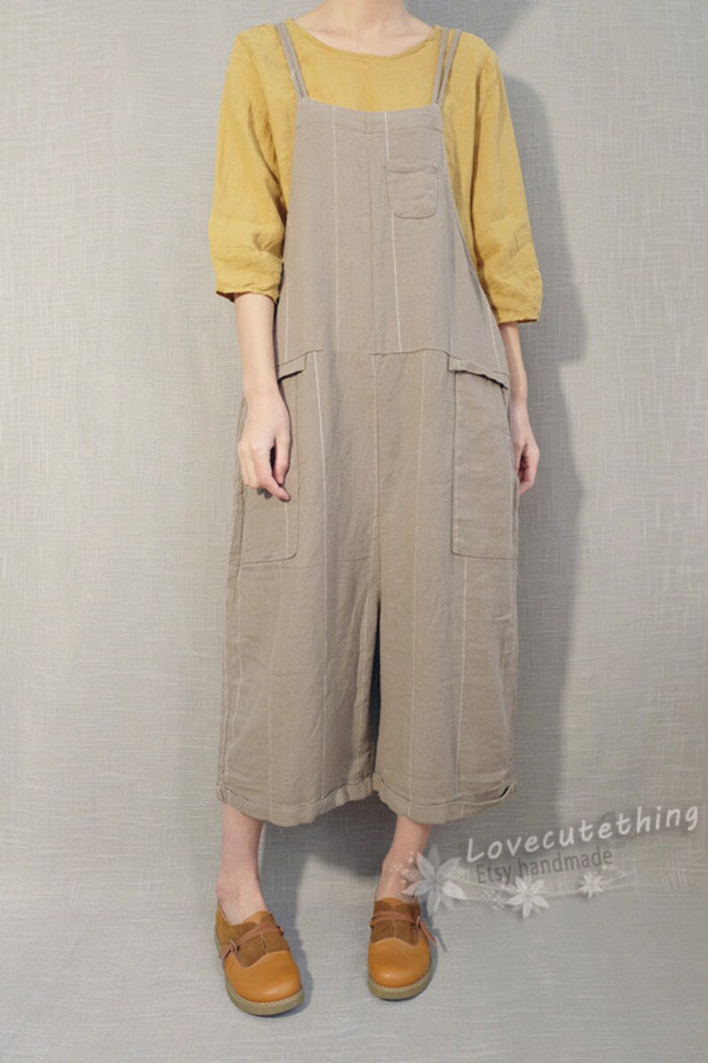 May include: A light brown linen overall dress with thin white stripes. The dress has two large pockets on the front and a smaller pocket on the chest. The model is wearing a yellow short-sleeved shirt under the dress and brown leather shoes.
