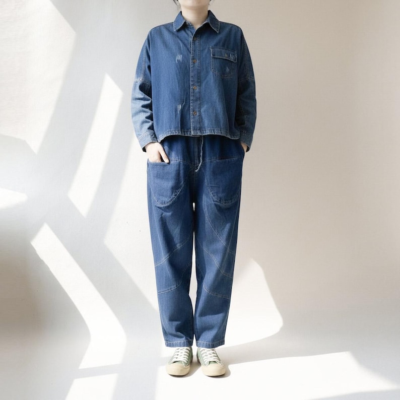 May include: A denim two-piece outfit, featuring a cropped button-down shirt and high-waisted pants with large pockets. The outfit is a medium blue wash, and the pants have a drawstring waist. The person is wearing light-colored sneakers.