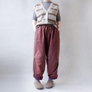 May include: A brown, loose-fitting pair of pants with a drawstring waist and a plaid trim at the cuffs. The pants are paired with a cream-colored knitted vest with a brown and white geometric pattern.