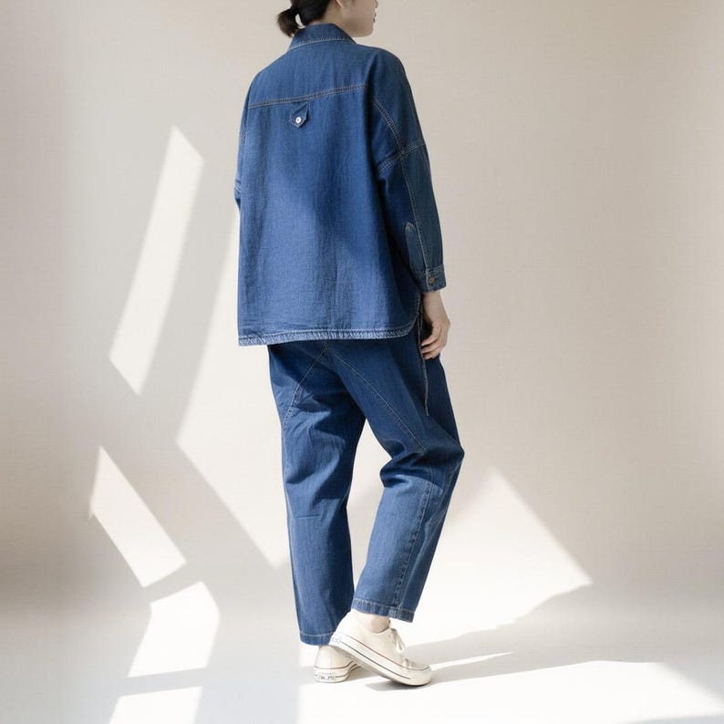 May include: A denim jacket and matching pants set in a medium blue wash. The jacket has a button closure and a single chest pocket. The pants are a relaxed fit. The outfit is paired with white canvas sneakers.