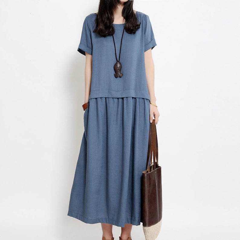 May include: A blue linen dress with a loose fit and a gathered skirt. The dress has short sleeves and a round neckline. The model is wearing a brown leather tote bag with a woven straw handle.