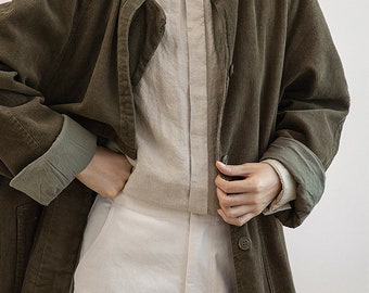 May include: A long, olive green corduroy jacket with a button-up front and a white collared shirt underneath.