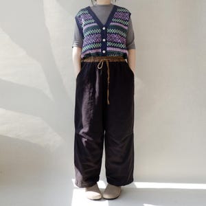 May include: A black sleeveless sweater vest with a geometric pattern in shades of purple, green, and blue. The vest has a button closure and is worn over a gray long-sleeved shirt. The person is wearing black wide-leg pants with a brown drawstring.