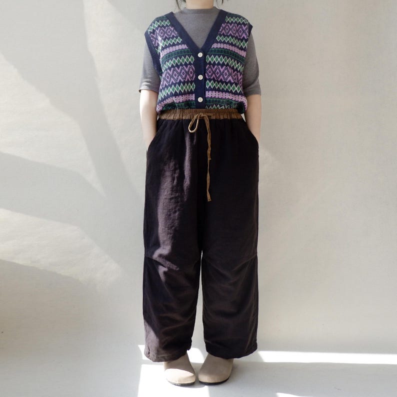 May include: A black sleeveless sweater vest with a geometric pattern in shades of purple, green, and blue. The vest has a button closure and is worn over a gray long-sleeved shirt. The person is wearing black wide-leg pants with a brown drawstring.