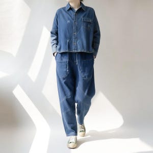 May include: A denim two-piece outfit. The top is a cropped button-down shirt with a chest pocket. The pants are loose-fitting with a drawstring waist and large front pockets. The outfit is a medium blue wash and is paired with canvas sneakers.