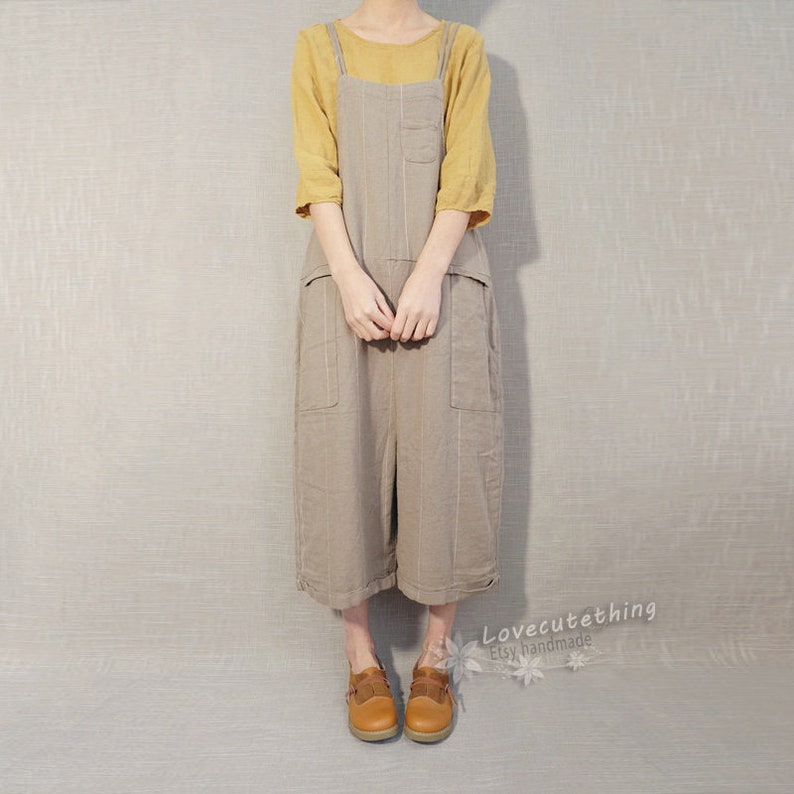 May include: A brown linen overall dress with thin vertical stripes and large pockets. The dress has adjustable straps and a relaxed fit. The model is wearing a yellow top underneath the dress.