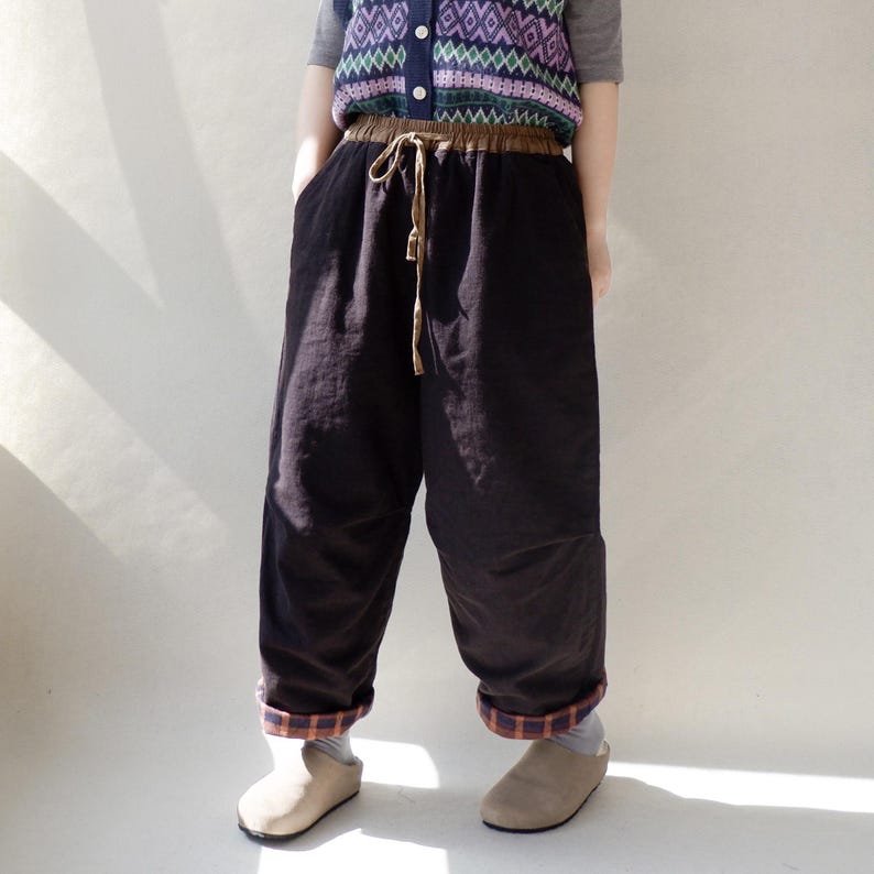 May include: Black wide-leg pants with a brown elastic waistband and a brown drawstring. The pants have a plaid trim at the bottom of the legs.