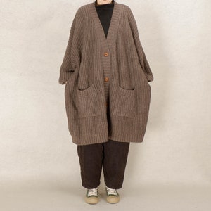 Op de afbeelding: Een lange bruine cardigan met een v-hals, knoopsluiting en twee klepzakken. De cardigan is gemaakt van een grof gebreid weefsel.