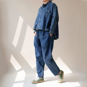 Könnte beinhalten: Ein zweiteiliges Jeans-Outfit, bestehend aus einem Hemd mit Knöpfen und einer passenden Hose. Das Hemd hat zwei Brusttaschen. Die Hose hat große Vordertaschen und eine lockere Passform. Das Outfit ist mittelblau und wird mit grünen Leinenschuhen getragen.
