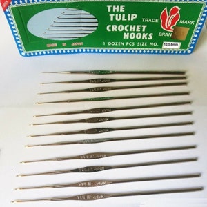 Könnte beinhalten: Ein Satz von 12 silbernen Häkelnadeln, beschriftet mit "TULIP JAPAN", in einer grün-weißen Schachtel. Die Schachtel trägt die Aufschrift "THE TULIP TRADE MARK CROCHET HOOKS 1 DOZEN PCS SIZE NO. 12/0.6mm".