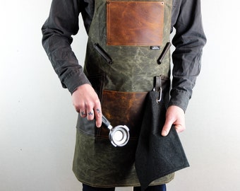 May include: A green canvas apron with brown leather accents and a black cloth towel hanging from a metal hook. The apron has a large pocket on the front and two smaller pockets on the sides. A person is holding a metal espresso tamper.