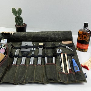 May include: A green canvas roll-up bag with leather accents and multiple pockets, containing a set of grilling tools, including a spatula, tongs, a brush, a knife, and a thermometer. The bag is rolled up and secured with a leather strap.