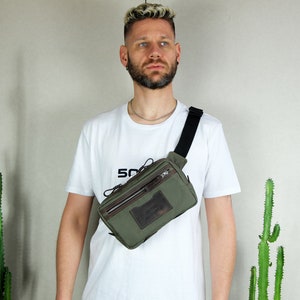 May include: A person wearing a white t-shirt with the word "SO" printed on it is wearing a green canvas fanny pack with a brown leather flap and zipper. The fanny pack is worn across the body.
