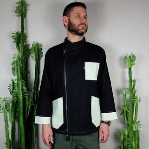 May include: A black chef jacket with a zip closure and contrasting light green panels. The jacket has a stand-up collar and two pockets on the front.