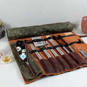 May include: A rolled-up leather and canvas grilling tool set with a variety of tools, including a spatula, tongs, a brush, and a knife. The set is brown and green and has a zippered pocket.