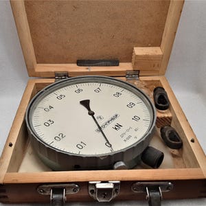 Könnte beinhalten: Ein mechanisches Messgerät mit weißem Zifferblatt, schwarzen Ziffern und einem Zeiger, untergebracht in einer offenen Holzkiste. Das Messgerät zeigt Zahlen von 0 bis 10 an, mit "KN" sichtbar. Die Kiste enthält auch zwei kleine, dunkle zylindrische Objekte.