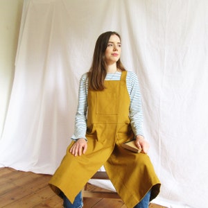 May include: Mustard-yellow apron with a large front pocket. The apron is worn over a striped long-sleeved top. The apron has a cross-back design and wraps around the body. The apron is made of a durable fabric.