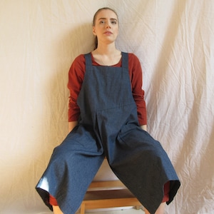 May include: A person wearing a dark blue denim overall dress with a red long-sleeved shirt underneath. The dress has two large pockets on the front and a wide leg.