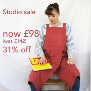May include: A woman wearing a red apron with a large pocket and a white and blue striped shirt is sitting on a chair. She is holding a yellow notebook. The text "Studio sale now £98 (was £142) 31% off" is visible in the image.