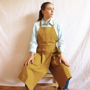 May include: Mustard-colored apron with a front pocket and a wrap-around design. The apron is made of a sturdy fabric and has a simple, functional design. The apron is worn over a light blue button-down shirt and blue jeans.