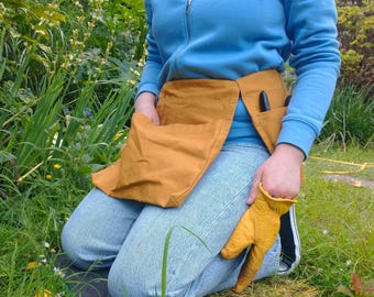 Cinturón utilitario tipo delantal de jardín: lona ocre con bolsillo grande y bolsillos para tijeras de podar y teléfono.