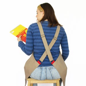 May include: A person wearing a tan apron with red straps and a blue striped shirt. The apron has crossed straps in the back. The person is holding a yellow and red spiral bound notebook with the text "9 to 5 DAILY ROUTINE" on the cover.