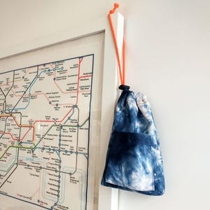 May include: A drawstring pouch featuring a blue and white tie-dye pattern with a front pocket and a bright orange cord. The pouch hangs from the corner of a white-framed map of the London Underground transit system on a plain white wall.