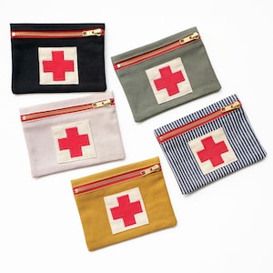 May include: Five small, rectangular pouches in various colours: black, olive green, light pink, mustard yellow, and blue and white striped. Each pouch features a red cross on a white square and a red zip.