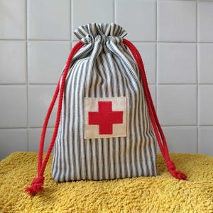 May include: A drawstring pouch made of white fabric with thin black vertical stripes. The bag features a square off-white patch on the front with a bold red cross symbol. Two thick red braided cords serve as the closure for the top of the bag.