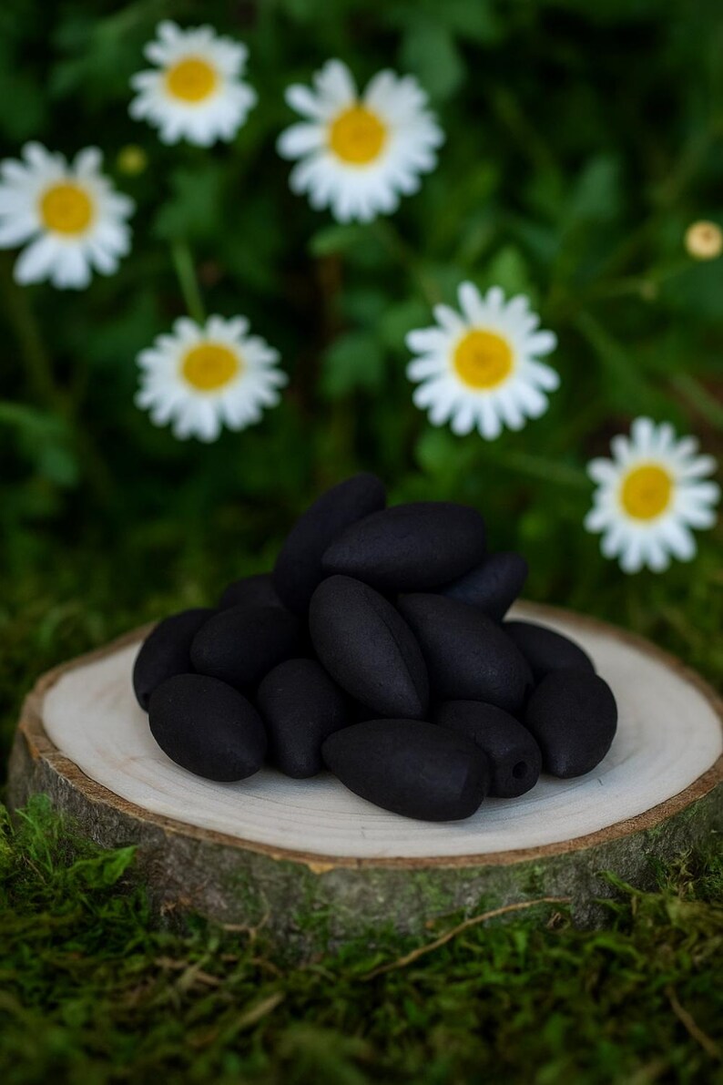 May include: A pile of black incense cones rests on a wooden slice, set against a backdrop of green foliage and white daisies with yellow centers. The cones have a smooth, matte finish.