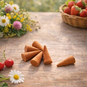 May include: Six brown incense cones are arranged on a wooden table, with a wicker basket of strawberries and wildflowers in the background. Two cherries and a daisy are in the foreground. The cones are for burning.