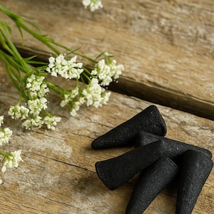 May include: A close-up shot of black incense cones arranged on a rustic wooden surface. Delicate white flowers with green stems are placed near the cones. The cones have a textured appearance.