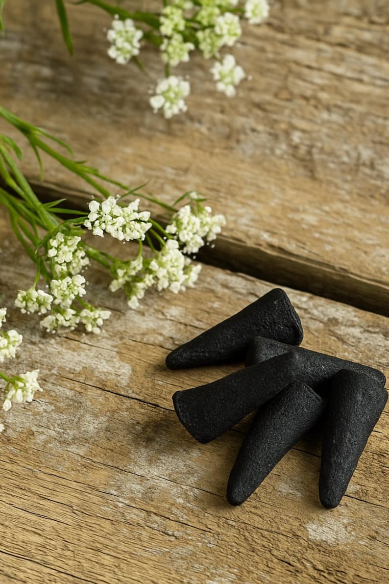 May include: A close-up shot of black incense cones arranged on a rustic wooden surface. Delicate white flowers with green stems are placed near the cones. The cones have a textured appearance.