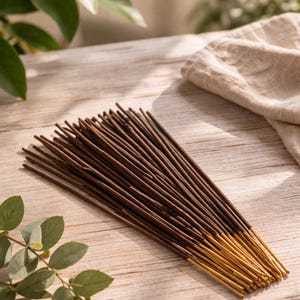 Op de afbeelding: Een bundel bruine wierookstokjes met goudkleurige uiteinden, gerangschikt op een licht houten oppervlak. Groene bladeren en een beige doek zijn op de achtergrond. De wierookstokjes zijn een natuurlijk product.
