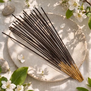 May include: A collection of dark brown incense sticks with gold tips, arranged on a light beige ceramic plate. The scene is decorated with white blossoms, green leaves, and small stones, creating a natural, serene aesthetic.