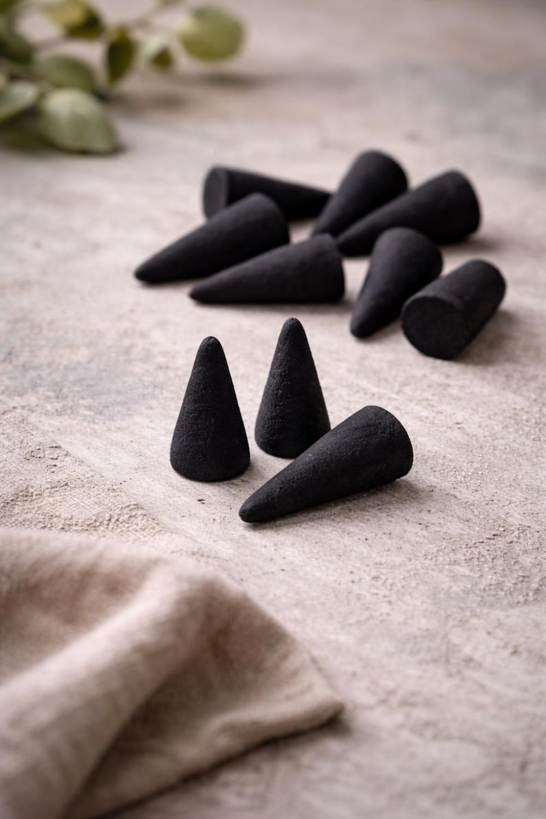 Op de afbeelding: Een verzameling zwarte wierookkegels gerangschikt op een getextureerd, lichtgekleurd oppervlak. De kegels staan in verschillende posities, sommige rechtop en andere op hun kant. Een gevouwen beige doek ligt op de voorgrond.
