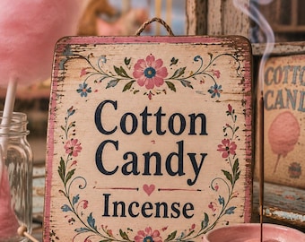 Może przedstawiać: Prostokątny drewniany szyld z postarzanymi krawędziami zawiera napis Cotton Candy Incense ciemnoniebieskimi literami, otoczony malowanymi różowymi i niebieskimi motywami kwiatowymi. Różowa miseczka zawiera palące się kadzidełko z dymem.