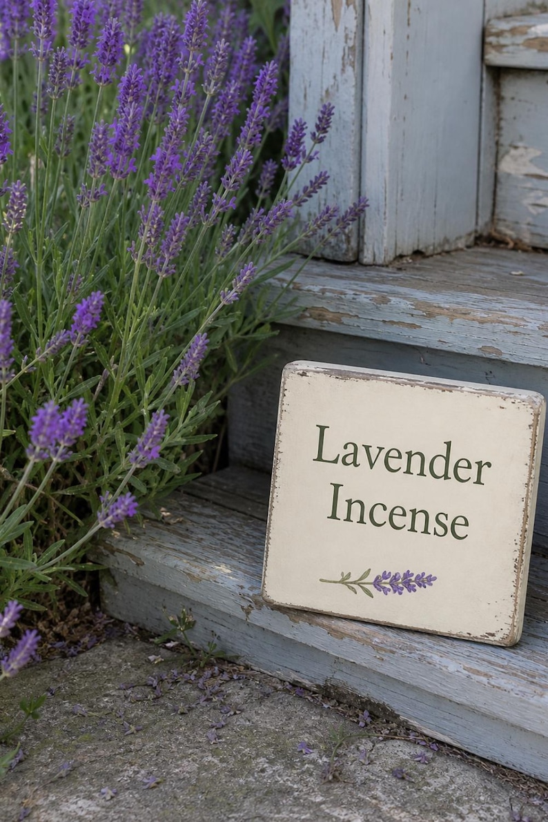 May include: A weathered, rectangular sign with the words "Lavender Incense" in dark green lettering, accompanied by a lavender sprig illustration. The sign rests on a rustic, light blue wooden surface, with vibrant purple lavender plants in the background.